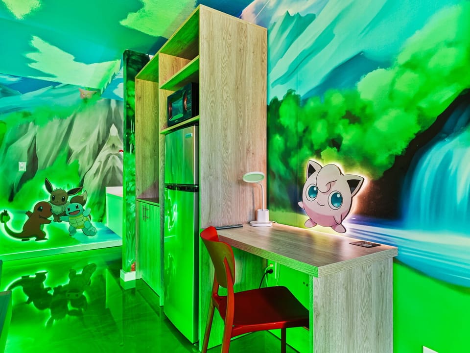 A colorful room featuring a desk, chair, and kitchenette with vibrant Pokémon-themed wall art.