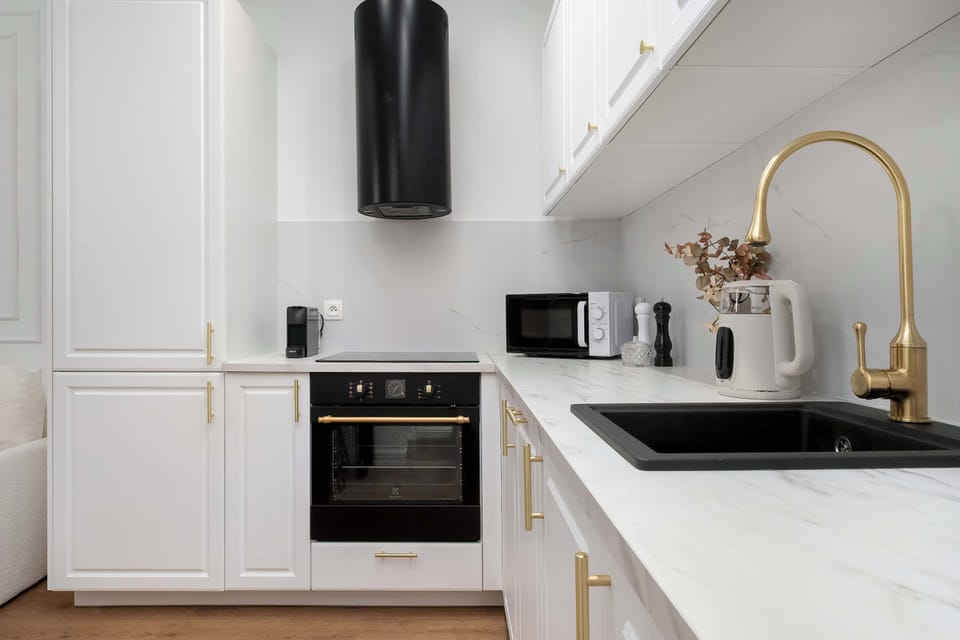 The kitchenette corner displays white cabinetry with golden handles and a black built-in oven under a matching hood. A black sink and modern appliances like a kettle and toaster complete this well-equipped culinary space.