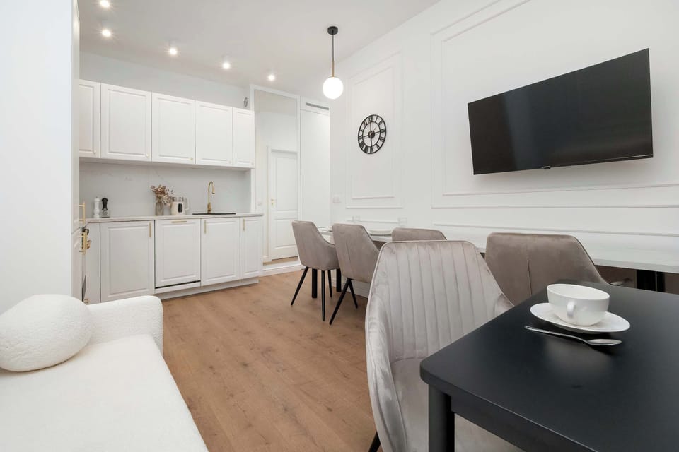 A functional kitchenette opens up to a dining space equipped with a black table and elegant upholstered chairs. A flat-screen TV is mounted on the wall, allowing guests to enjoy entertainment while cooking or eating.