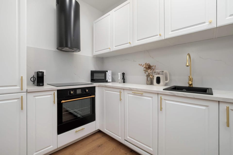 An L-shaped white kitchenette features a clean, bright aesthetic with a contrasting black countertop and gold accents. The design is both stylish and practical, offering plenty of storage for short-term stays.