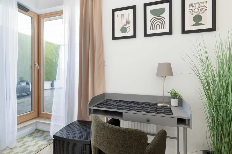 A compact workspace by a window with a desk and chair. Framed pictures and a tall plant add warmth to the setting.