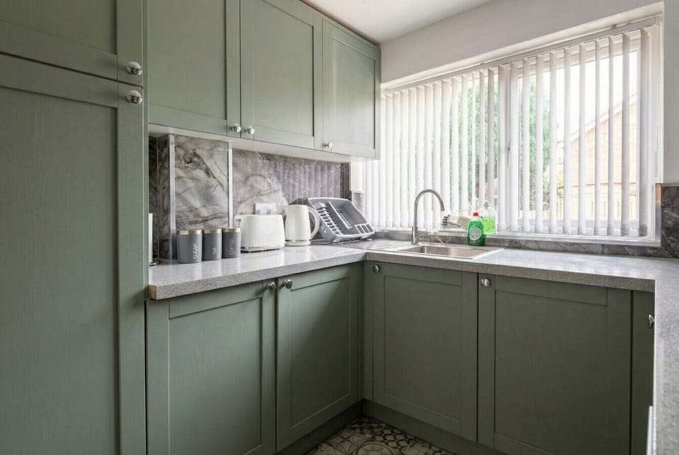 Sage-green kitchen with stone worktop, stainless sink, a kettle, and patterned tile flooring beside a window.
