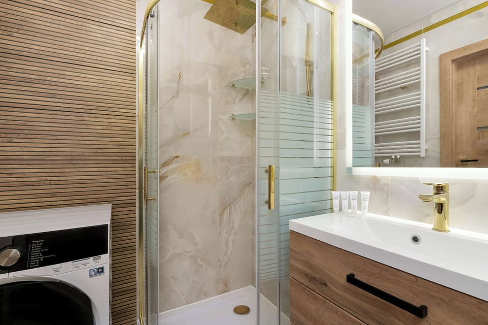 The bathroom is designed with luxury in mind, featuring marble-effect tiles and a spacious walk-in shower with a gold-finished rain shower head. A wooden vanity with a white ceramic sink sits under a well-lit mirror.