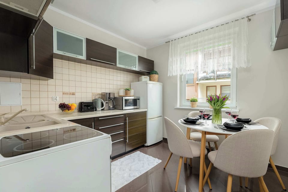 A functional kitchen area combines dark wood cabinetry with white upper units, featuring a dining table with upholstered chairs and a bright window.