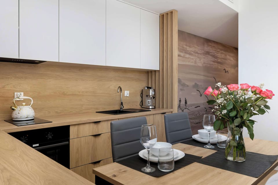 A compact kitchenette with light wood cabinets, white upper units, and a clean countertop. Decorative items and a vase of flowers bring warmth to the minimalist design.