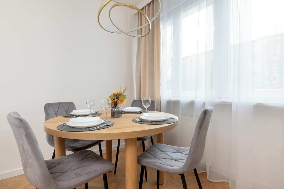 A dining area with a small round table and two upholstered chairs. A simple decorative centerpiece rests on the tabletop. Light streams in from a nearby window, brightening the space.
