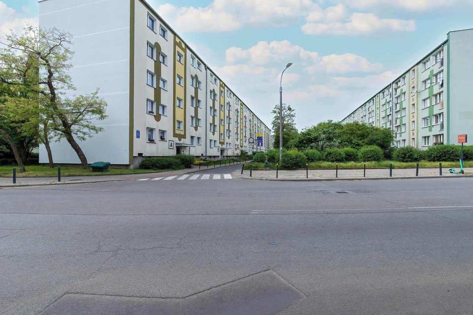Another exterior shot of a long apartment building with parked cars in front. The sky is partly cloudy, and greenery lines the property. The residential complex appears quiet and well-maintained.