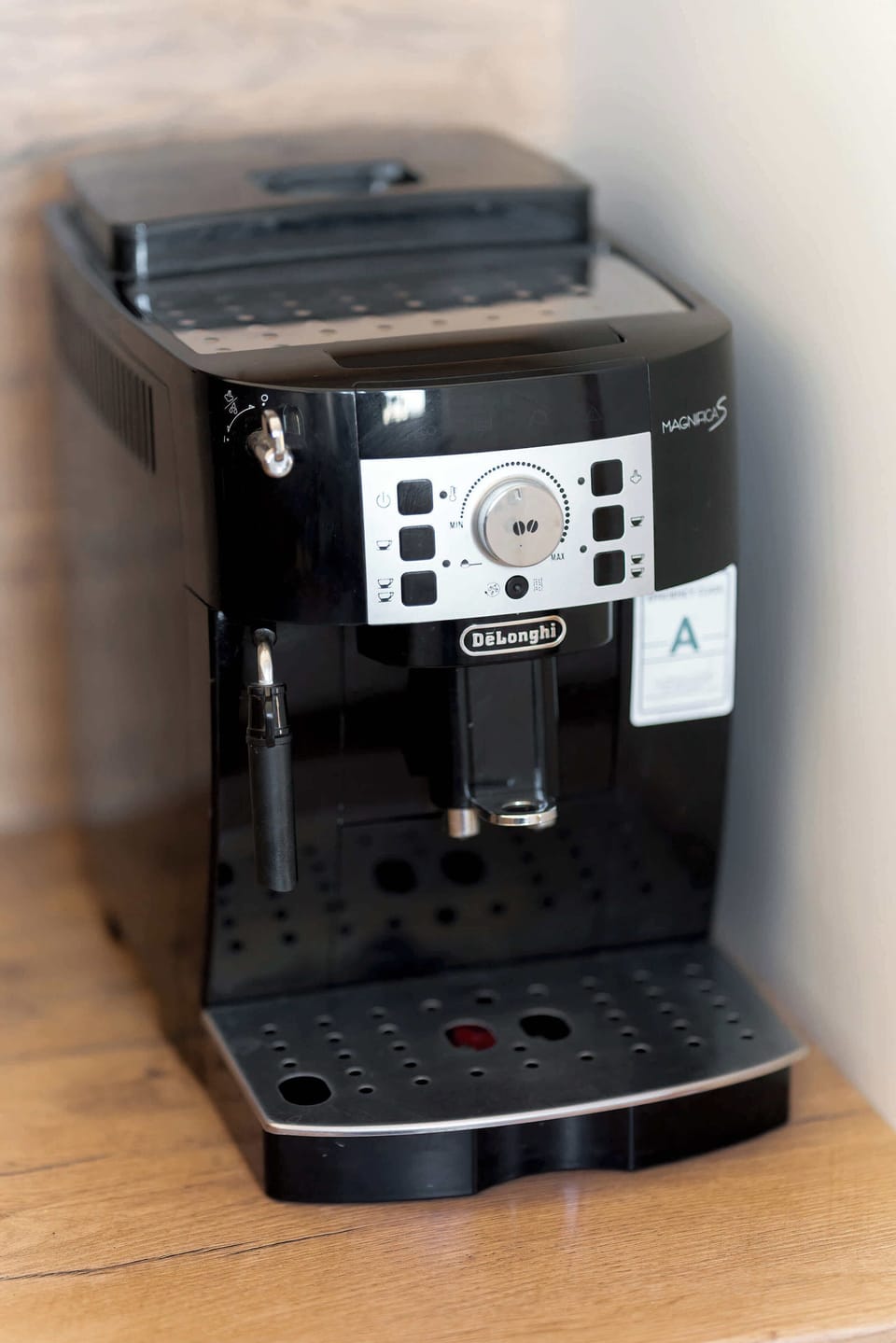 A close-up of a coffee machine placed on a kitchen counter. The machine stands on a wooden surface with simple kitchen décor nearby. The appliance suggests convenience for preparing hot drinks.