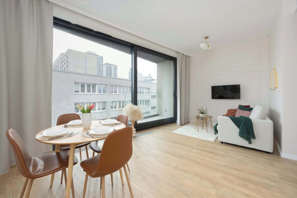 A dining area positioned beside a wide floor-to-ceiling window. A round table with upholstered chairs overlooks city buildings. Natural light fills the space generously.