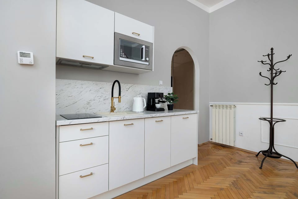 A minimalist white kitchenette showing handleless cabinetry and a white marble-patterned backsplash. The space is equipped with built-in appliances and a stylish gold-accented faucet.