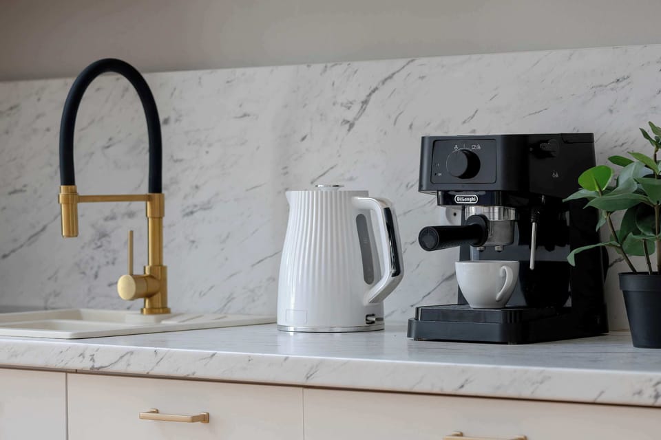 A kitchenette countertop detail highlighting a modern espresso machine and a white electric kettle. The gold faucet and marble backsplash add a luxurious feel to the workspace.