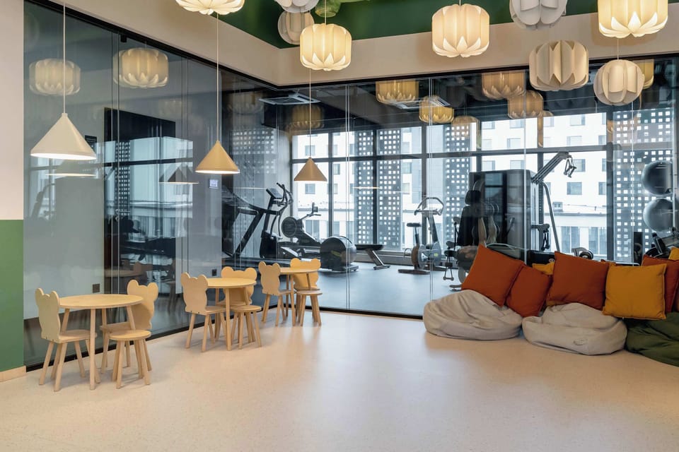 Children's playroom with chairs and cushions for playing and gym area with exercise machines and large windows. Natural light brightens the fitness space. The equipment looks modern and well maintained.