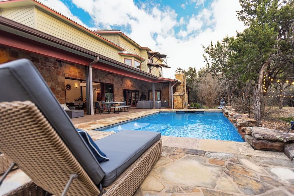 Modern luxury home featuring a stunning pool area surrounded by mature trees and natural stone landscaping under bright blue skies.