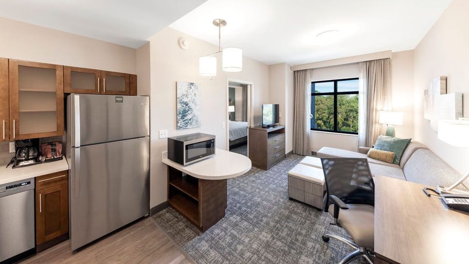 The open living area combines a sofa bed, desk with chair, kitchen area, and a window providing natural light.