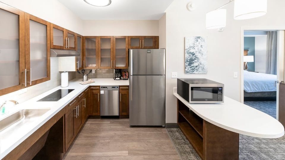 The kitchen features stainless steel appliances, wooden cabinets, a sink, and a stovetop set into a light-colored countertop.