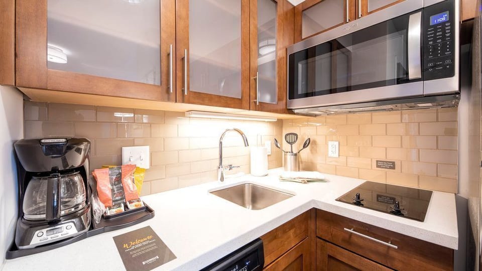 A kitchen corner with a coffee machine, a sink, a microwave, and organized cabinets with utensils.