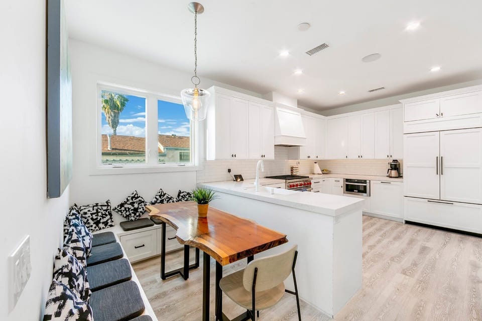 A functional and stylish dining area featuring a custom built-in banquette with navy patterned upholstery. The adjacent kitchen boasts a large waterfall-edge island, high-end white cabinetry, and designer pendant lighting.