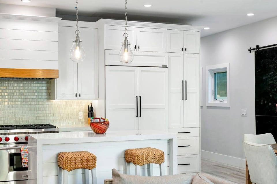 A stunning view of the chef-inspired kitchen, featuring a large white waterfall-edge island paired with three stylish woven barstools. Professional-grade stainless steel appliances and custom cabinetry create a high-end culinary environment.