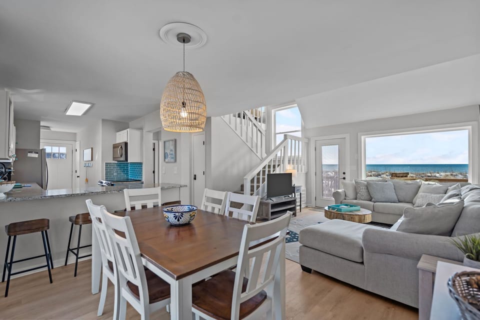 Bright open-concept space in Oak Island NC featuring wood dining table, kitchen with granite counters and blue tile backsplash, breakfast bar, sectional sofa, and ocean views through large windows and deck door.