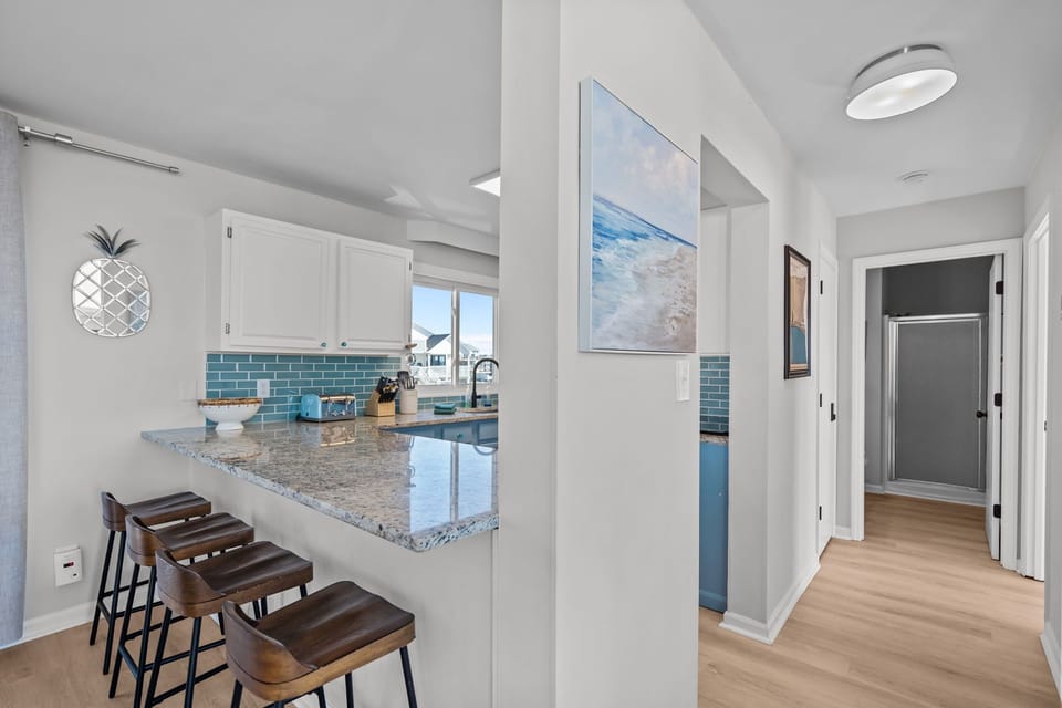 Bright coastal kitchen in Oak Island NC featuring granite countertops, breakfast bar with stools, stainless appliances, blue subway tile backsplash, window over sink, and full-size washer and dryer nearby.