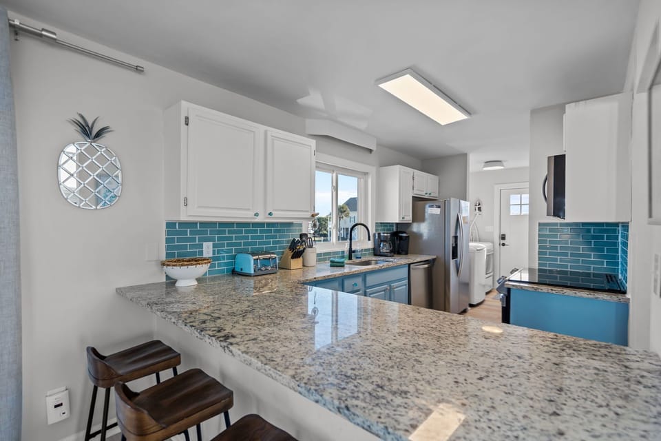 Bright coastal kitchen in Oak Island NC featuring granite countertops, breakfast bar with stools, stainless appliances, blue subway tile backsplash, window over sink, and full-size washer and dryer nearby.