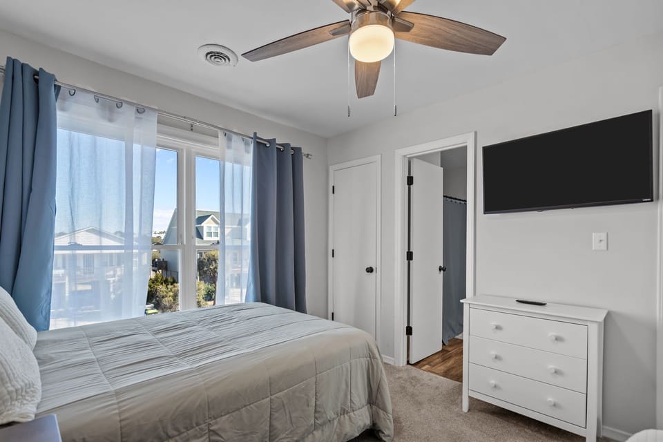 Cozy Oak Island NC bedroom with queen and twin beds, soft neutral bedding, nightstand with lamp, ceiling fan, octopus artwork, carpeted floors, and coastal décor for a relaxing beach retreat.