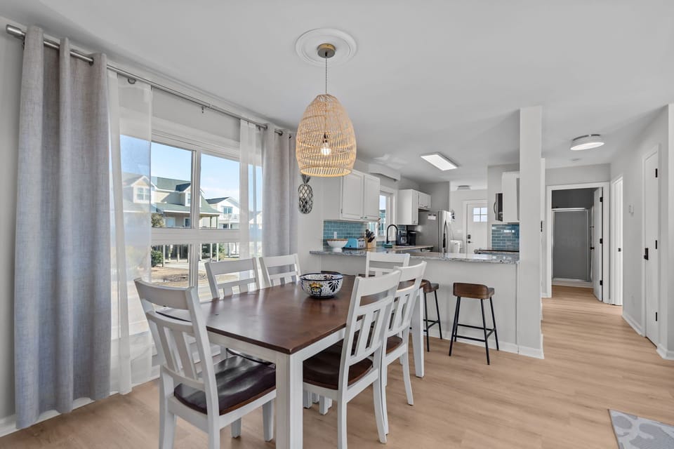 Bright open-concept space in Oak Island NC featuring wood dining table, kitchen with granite counters and blue tile backsplash, breakfast bar, sectional sofa, and ocean views through large windows and deck door.