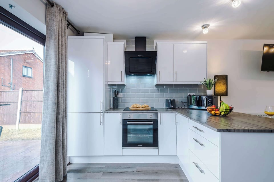 Modern kitchen with sleek white cabinets, built-in oven, electric stovetop, microwave, toaster, coffee machine, and knife block. Stylish black range hood and light gray tile backsplash complete the clean, contemporary look.