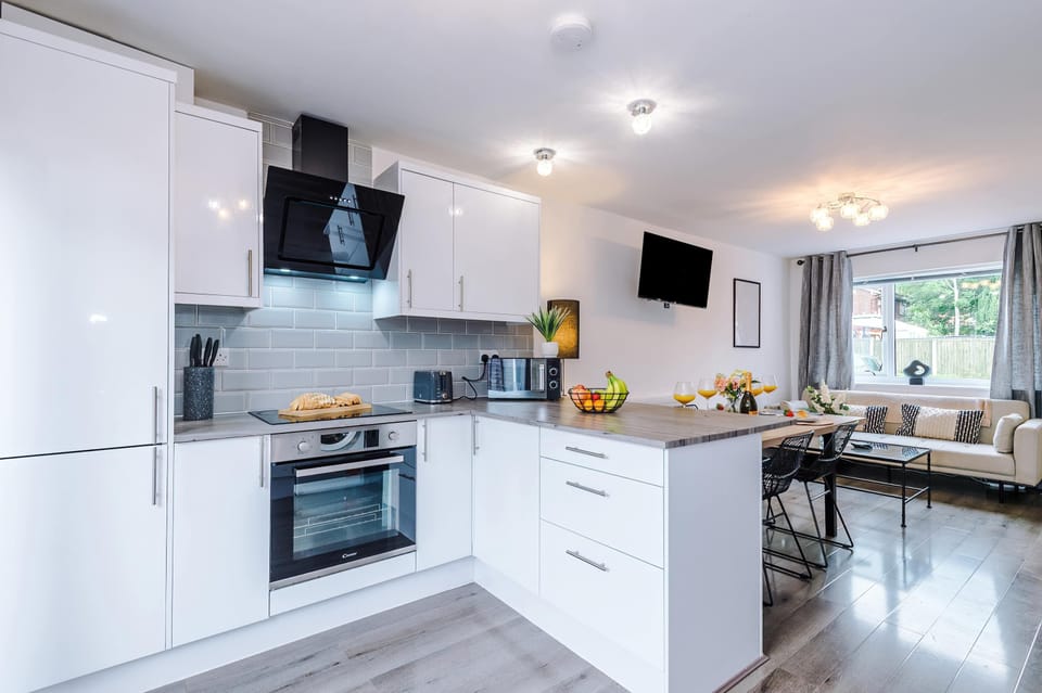 Modern open-plan kitchen and living area with sleek white cabinets, built-in oven, black glass cooktop, microwave, toaster, and stylish range hood. Includes cozy lounge, dining table, and wall-mounted TV in a bright, welcoming space.