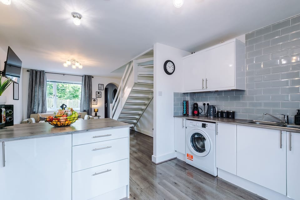 Contemporary open-plan kitchen and living area with white cabinetry, stainless steel appliances including washing machine, built-in oven, microwave, and sleek cooktop. Features stylish gray tile backsplash, wall-mounted TV, and staircase to upper lev