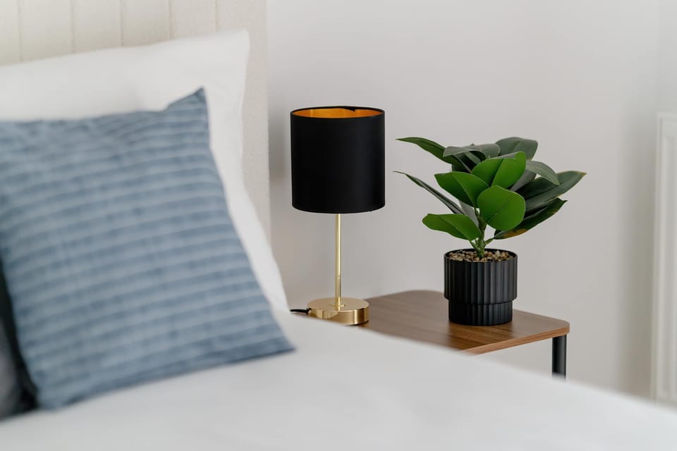 A close-up shot focuses on a stylish bedside setup, featuring a dark lamp with a golden interior and a small green plant. The soft textures of the pillow and bedding suggest a high level of comfort.