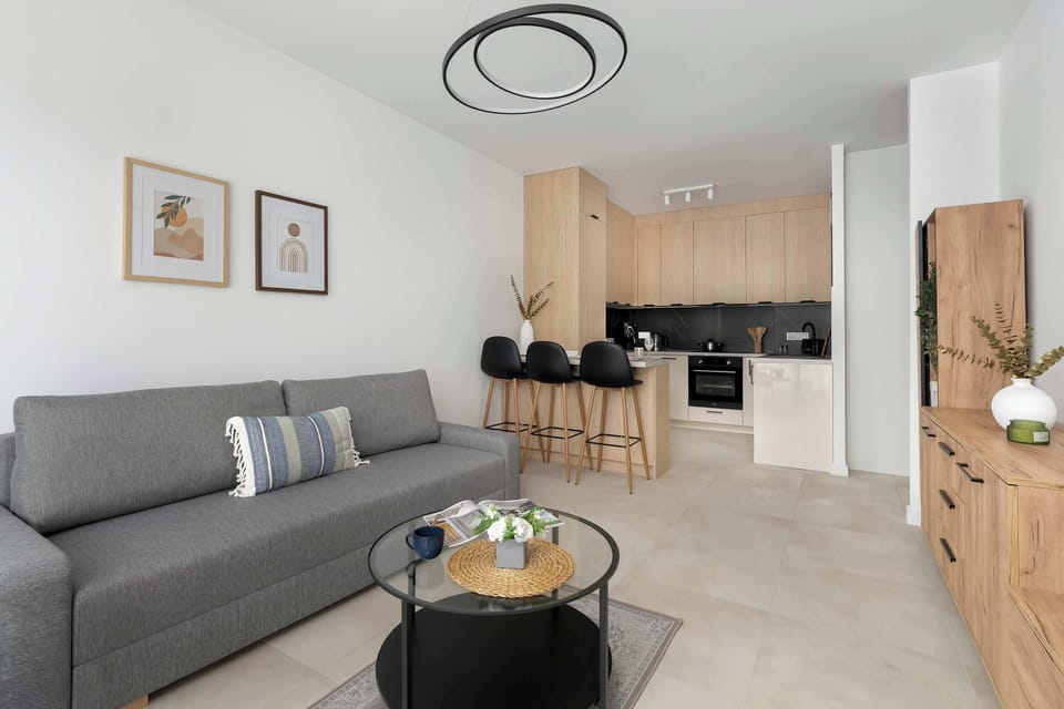 The open-concept living area combines a comfortable grey sofa with a modern wooden kitchenette in the background. A round black coffee table sits on a light-colored floor, creating a functional relaxation space.
