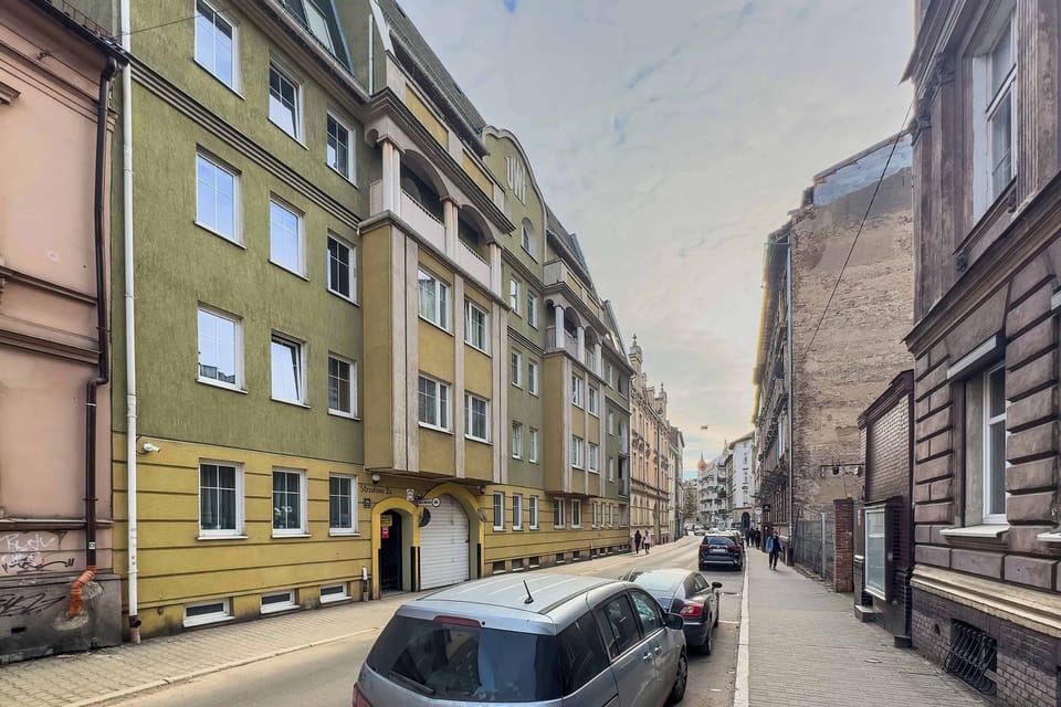 A street-level view of a row of colorful residential buildings along a narrow city street with parked cars lining the road.