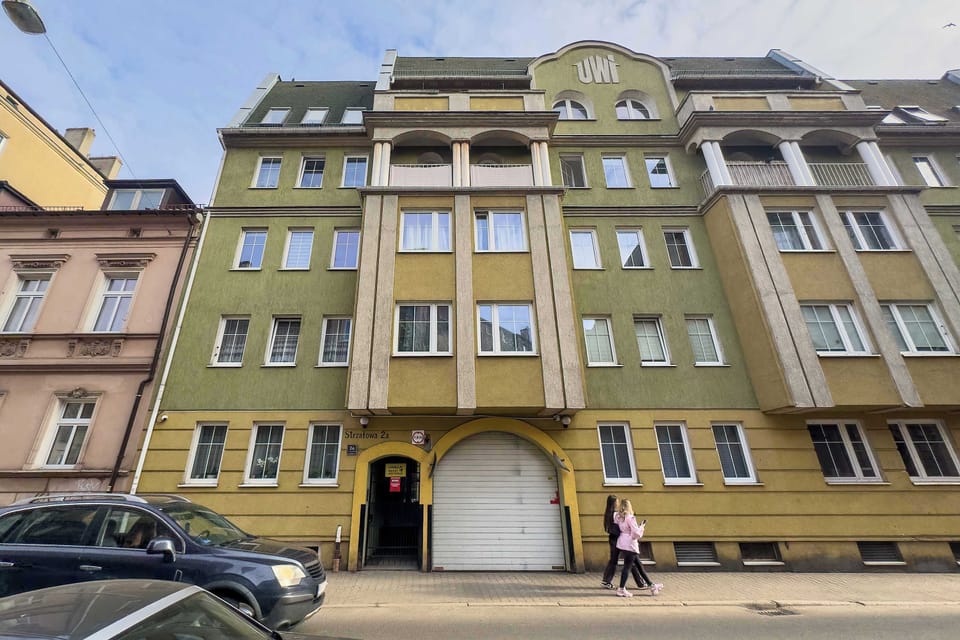 The exterior of a multi-story residential building painted in muted green with decorative window frames and parked cars in front.