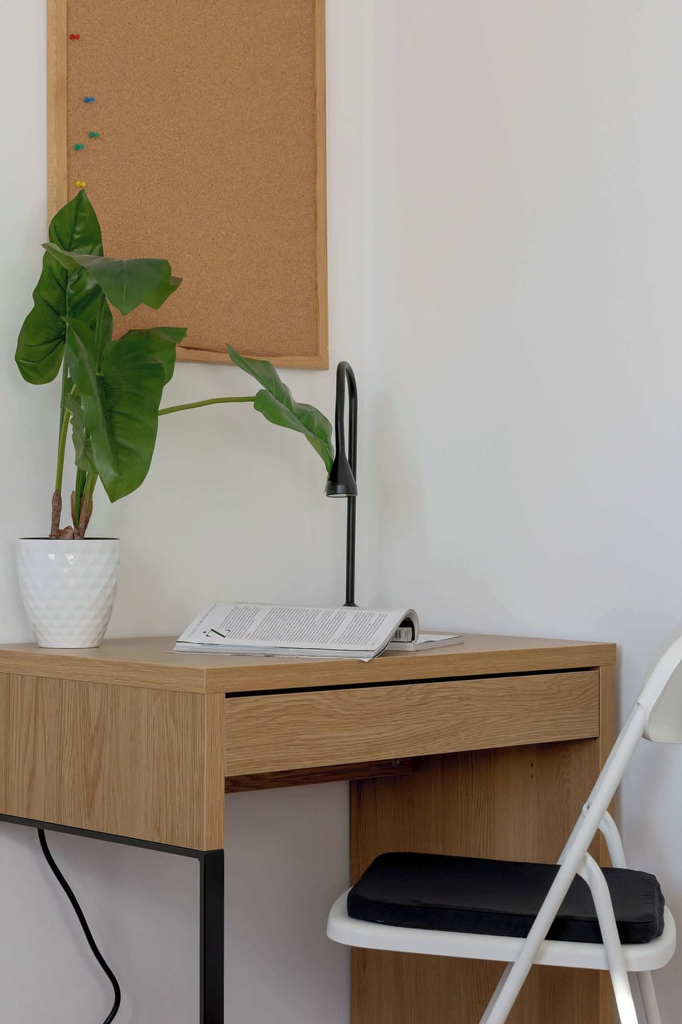 A simple light-wood desk with a small stool and a potted green plant stands against a white wall next to a tall wooden shelf.