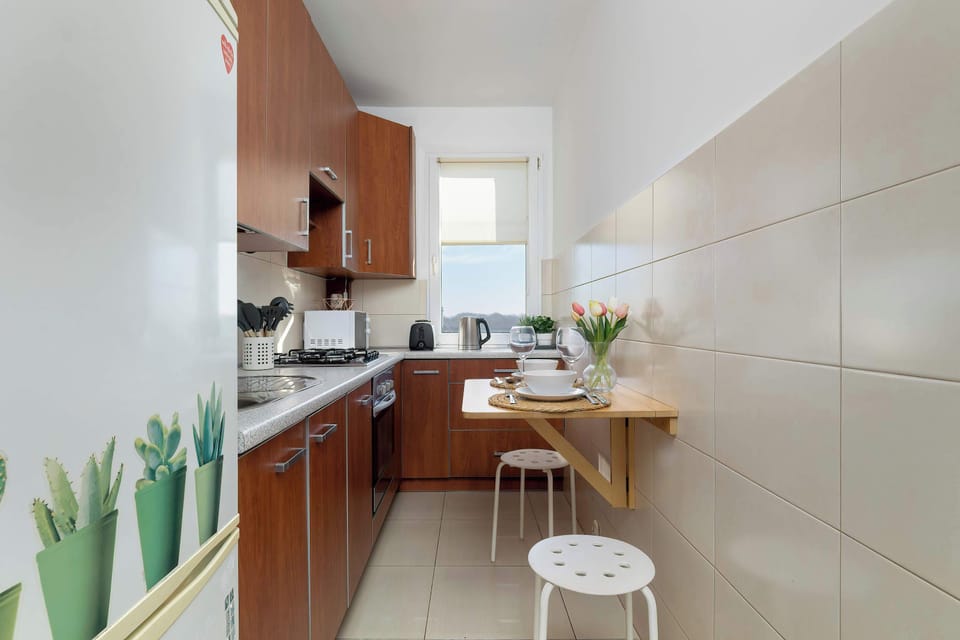 A narrow kitchen space with a small breakfast bar and two stools, white tile walls, and a large green potted plant in the corner.