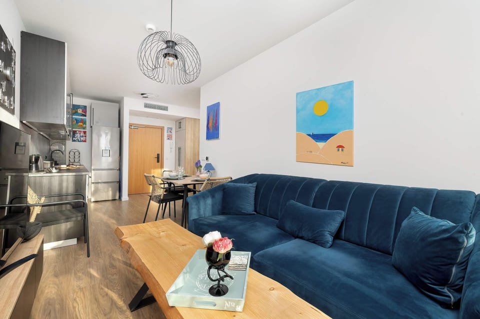 An open-plan living space that seamlessly connects the lounge area to the kitchen. The blue sofa serves as a pop of color against the white walls and light wood floors.