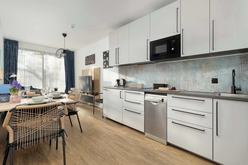 A clean white kitchenette equipped with modern appliances and light grey cabinetry. A dining area with a wooden table is positioned nearby, making the most of the open-plan layout.