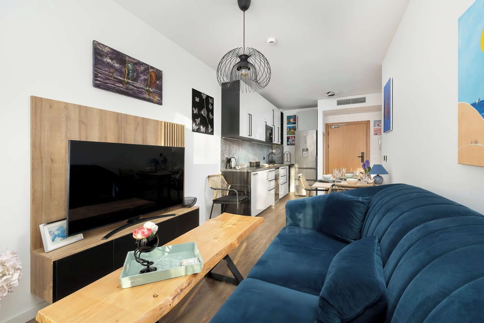 A view of the living room from the dining table, showing the TV area and a colorful beach-themed painting. The layout is designed for both relaxation and entertaining.