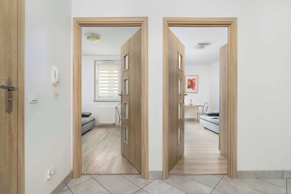 A hallway with two sets of wooden doors leads to different parts of the apartment. An open view through a doorway shows a window, which helps illuminate the corridor.