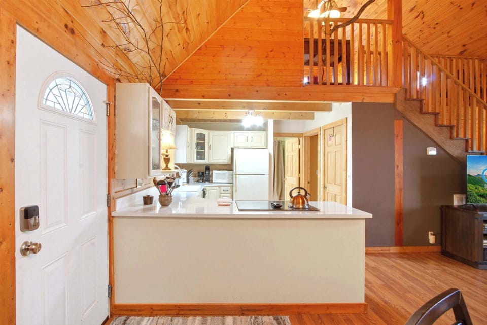 A clean, open-concept kitchen featuring cream-colored cabinetry, a white countertop with an integrated electric cooktop, and a double-basin sink situated beneath a window with forest views.
