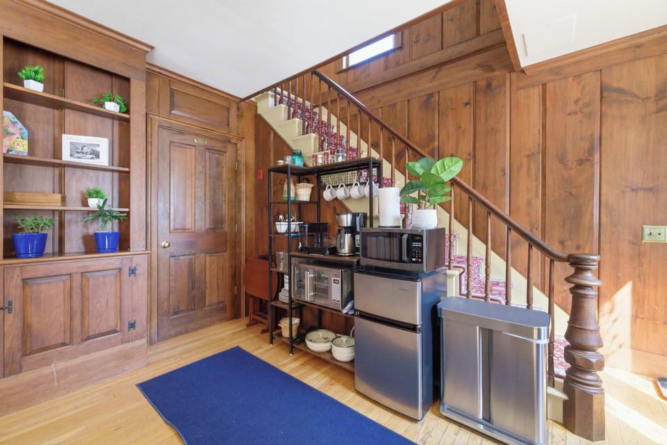 The kitchenette and private entrance from a wider angle, showing the full setup under the staircase with the blue entry mat and wood-paneled walls.