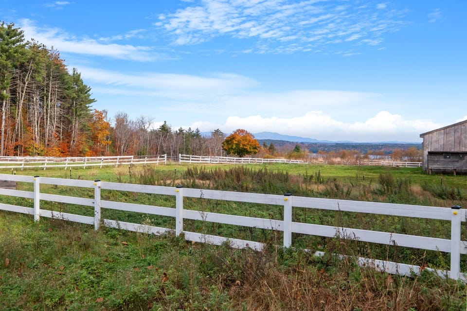 White post-and-rail fencing borders the farm fields in fall, with the mountains visible on the horizon and the historic barn in the background.