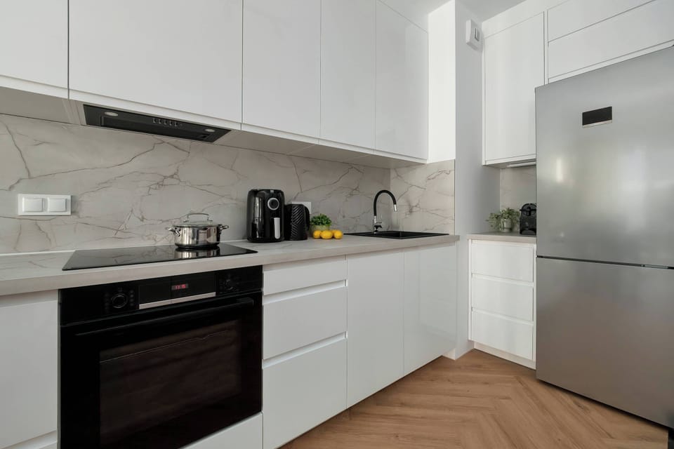 This picture shows a white kitchen with modern appliances. Wooden pattern can be seen in the flooring, while on the right there is a large silver refrigerator.