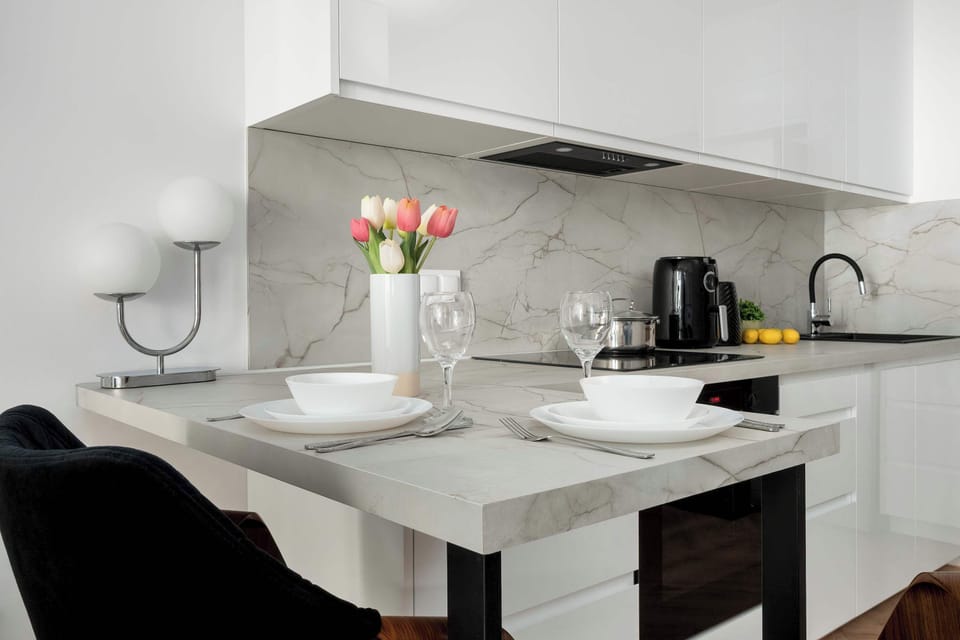 A modern kitchen with a large dining island that can also function as a breakfast bar. There are two black bar stools on one side and a white flower arrangement with other dishes on top of the counter.