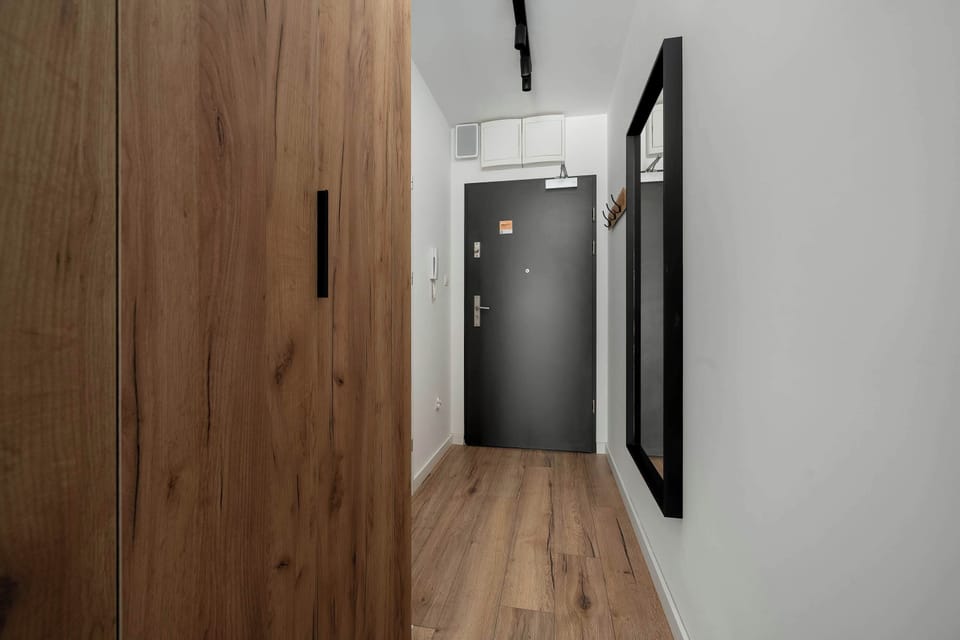 A hallway with wooden wardrobe doors on one side and a dark entrance door at the end. Light walls and flooring keep the corridor bright. The layout suggests storage space near the apartment entrance.
