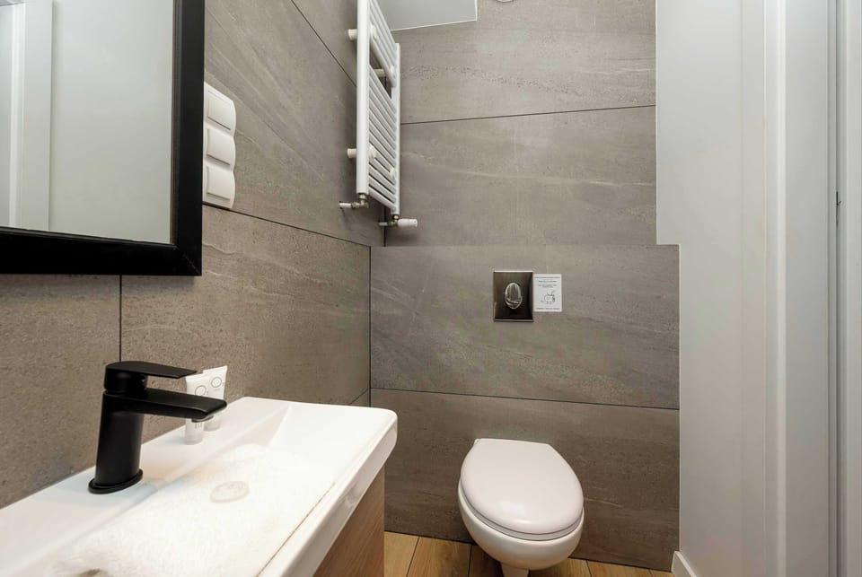 A bathroom corner with a toilet and sink positioned close together. A heated towel rack is mounted on the wall above the toilet. Light-colored tiles make the room appear bright and tidy.