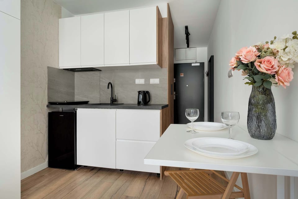 A wider view of the kitchenette area with white cabinets, a sink, and small appliances on the counter. A small round dining table with wooden chairs stands nearby. The space is compact but efficiently arranged.
