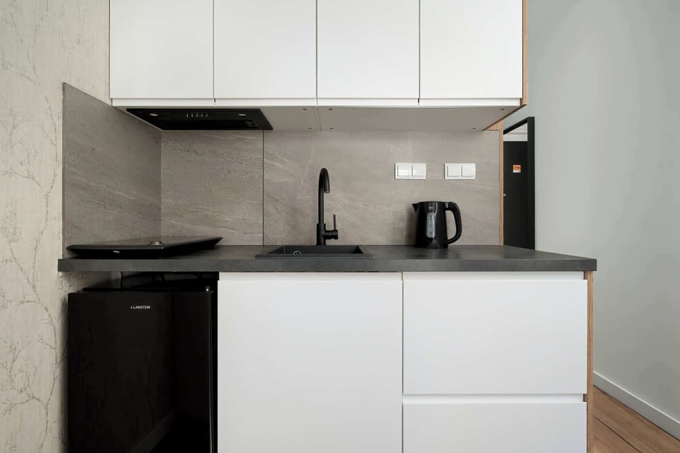 A modern kitchenette with white upper cabinets and darker lower cabinets. A black countertop holds small appliances such as a kettle. The backsplash and clean lines give the kitchen a sleek, contemporary look.