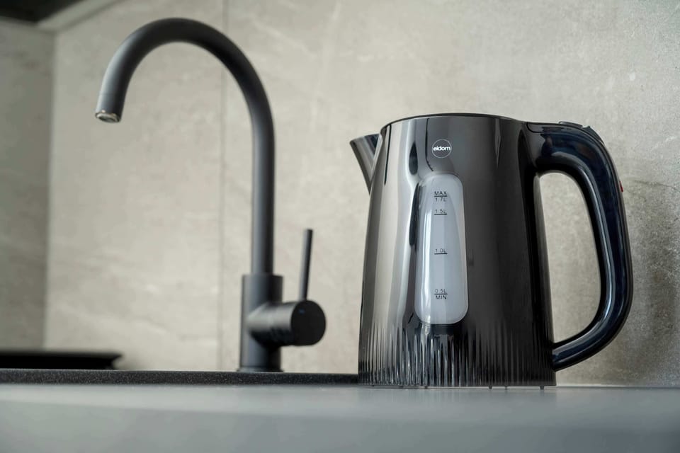A close-up of a kitchen counter featuring a black faucet and an electric kettle. Light-colored wall tiles form the backsplash behind the sink area. The setup looks simple, functional, and modern.